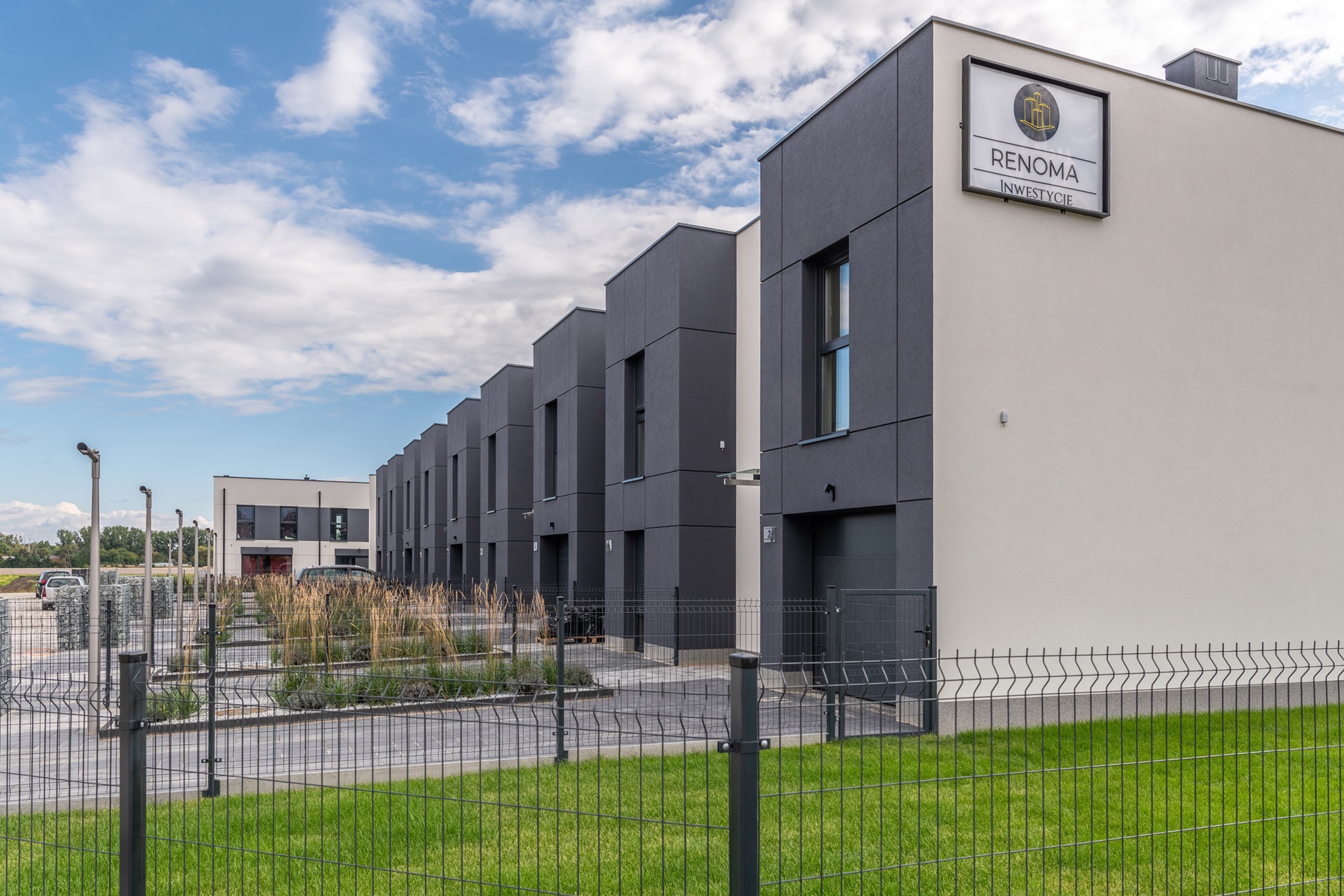 Szereg nowoczesnych, dwukondygnacyjnych domów z płaskimi dachami, wykończonych w odcieniach szarości i bieli, ogrodzonych metalowym płotem na tle błękitnego nieba.