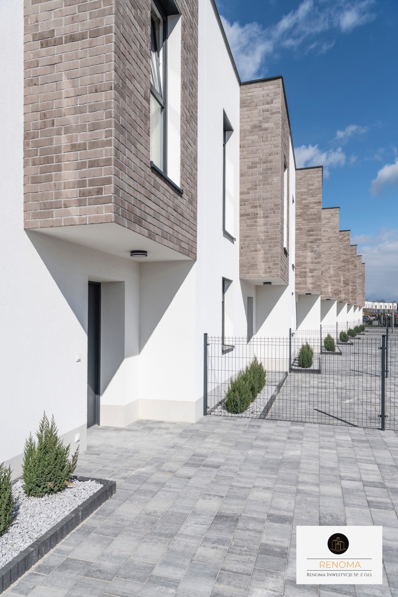 Szereg identycznych, nowoczesnych domów z białymi ścianami i ceglanymi elementami, widok z zewnątrz, z kostką brukową i niskimi krzewami wzdłuż ogrodzenia.