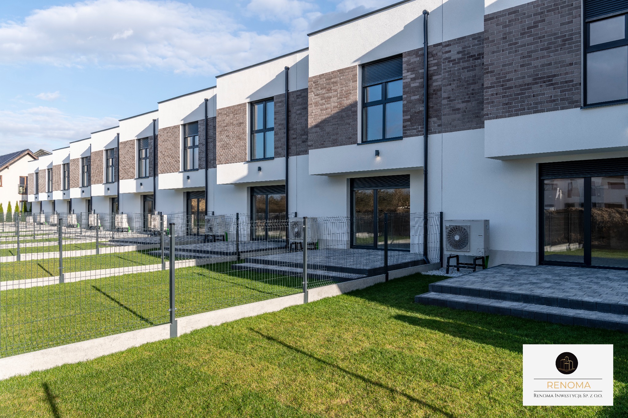 Szereg domów jednorodzinnych z ceglanymi elementami elewacji, ogrodzonych siatką, z widocznymi klimatyzatorami na zewnątrz. Nowoczesna architektura, zadbany trawnik.