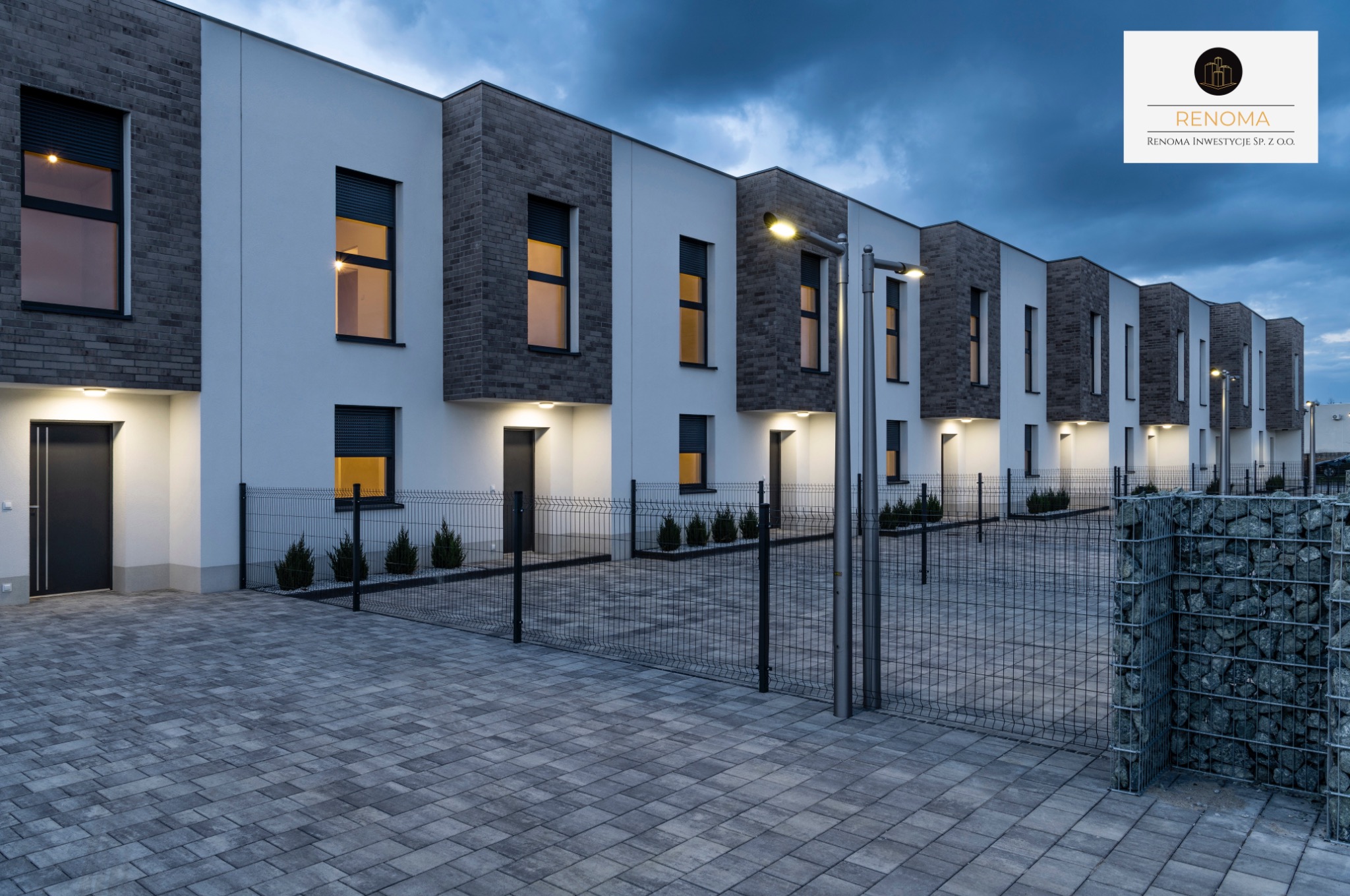 Szereg nowoczesnych domów jednorodzinnych z elementami z cegły, oświetlonych lampami ulicznymi, ogrodzonych siatką, z kostką brukową na podjeździe, nocne ujęcie.