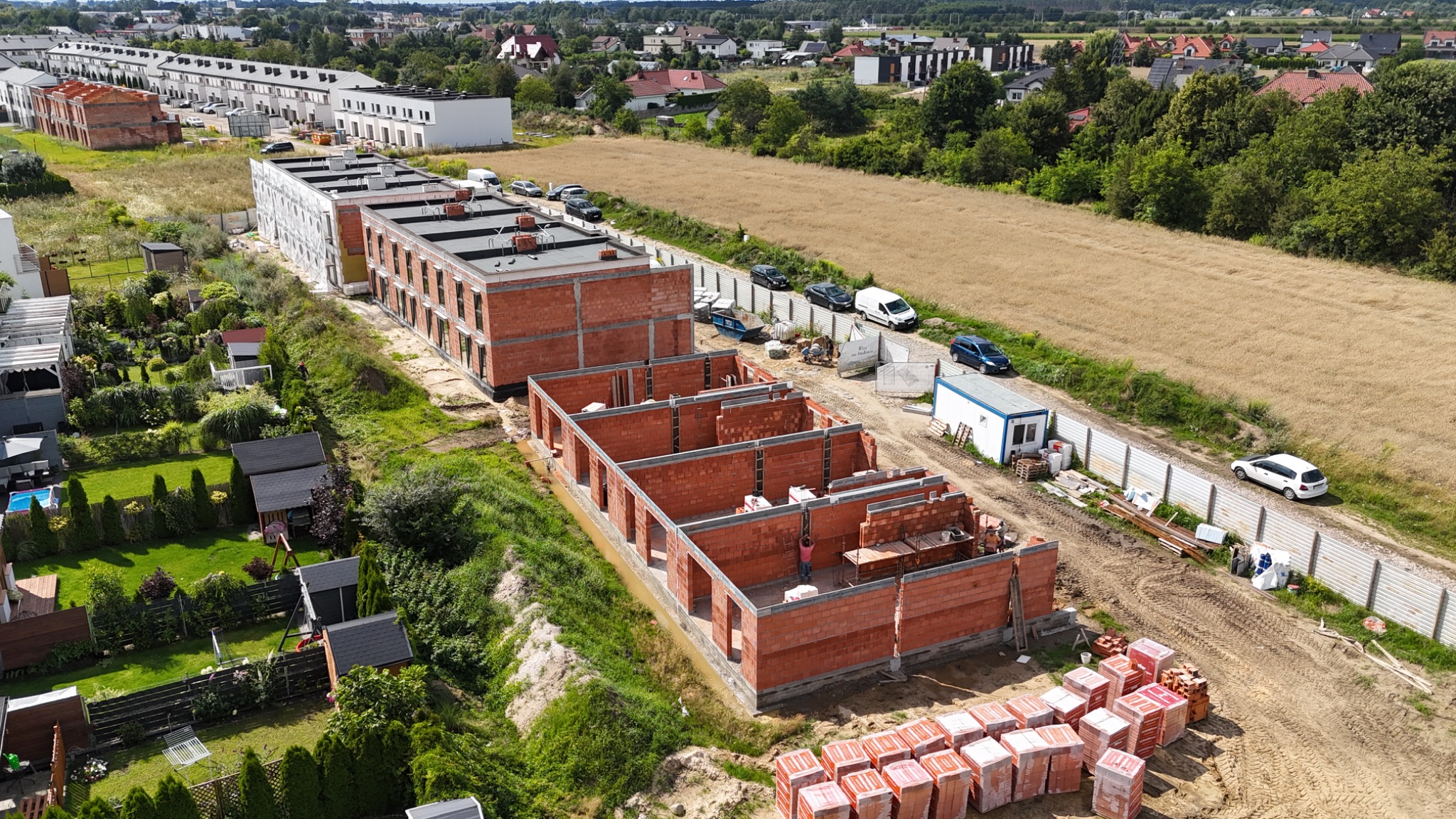 Budowa osiedla domów z czerwonej cegły, widok z góry. Etap murowania ścian, widoczne palety z cegłami i pracownik wewnątrz konstrukcji.