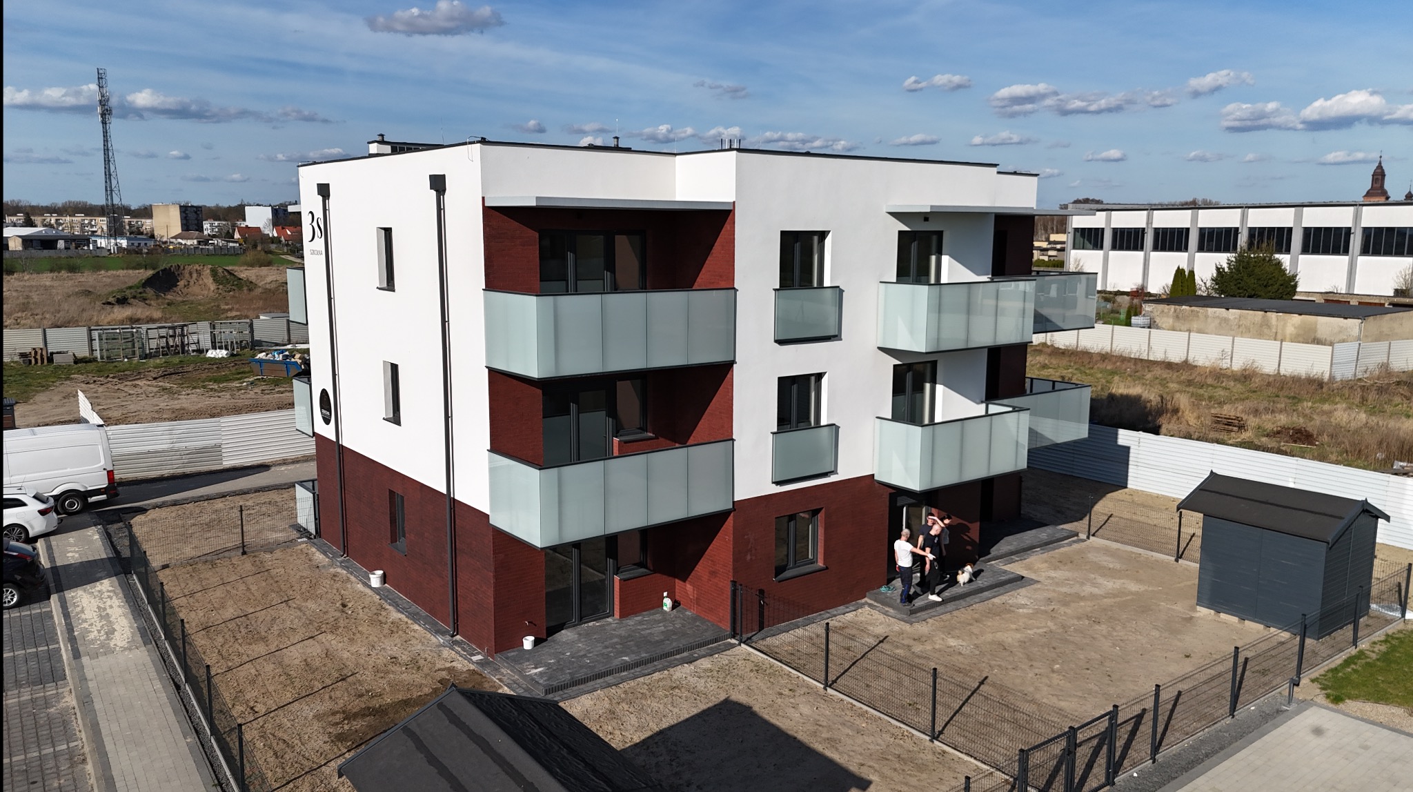 Nowoczesny, trzypiętrowy budynek z balkonami, wykończony cegłą i białym tynkiem. Widoczne osoby na parterze, ogrodzenie i otoczenie w trakcie zagospodarowywania.