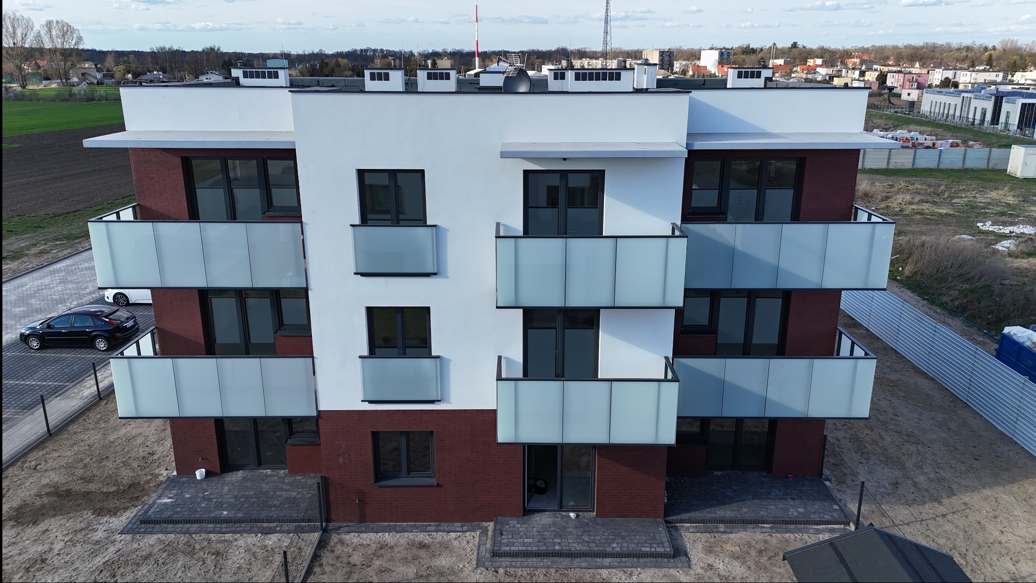 Nowoczesny, trzypiętrowy budynek z balkonami o szklanych balustradach, wykończony cegłą i białym tynkiem. Widok z góry, w tle zabudowa miejska i pole.