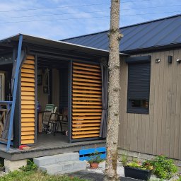 P.K-Woods. - Nowoczesna, drewniana altana z przesuwnymi żaluzjami, szarą zasłoną i ciemnym dachem. Widoczne rośliny w donicach i fragment pnia drzewa na pierwszym planie.