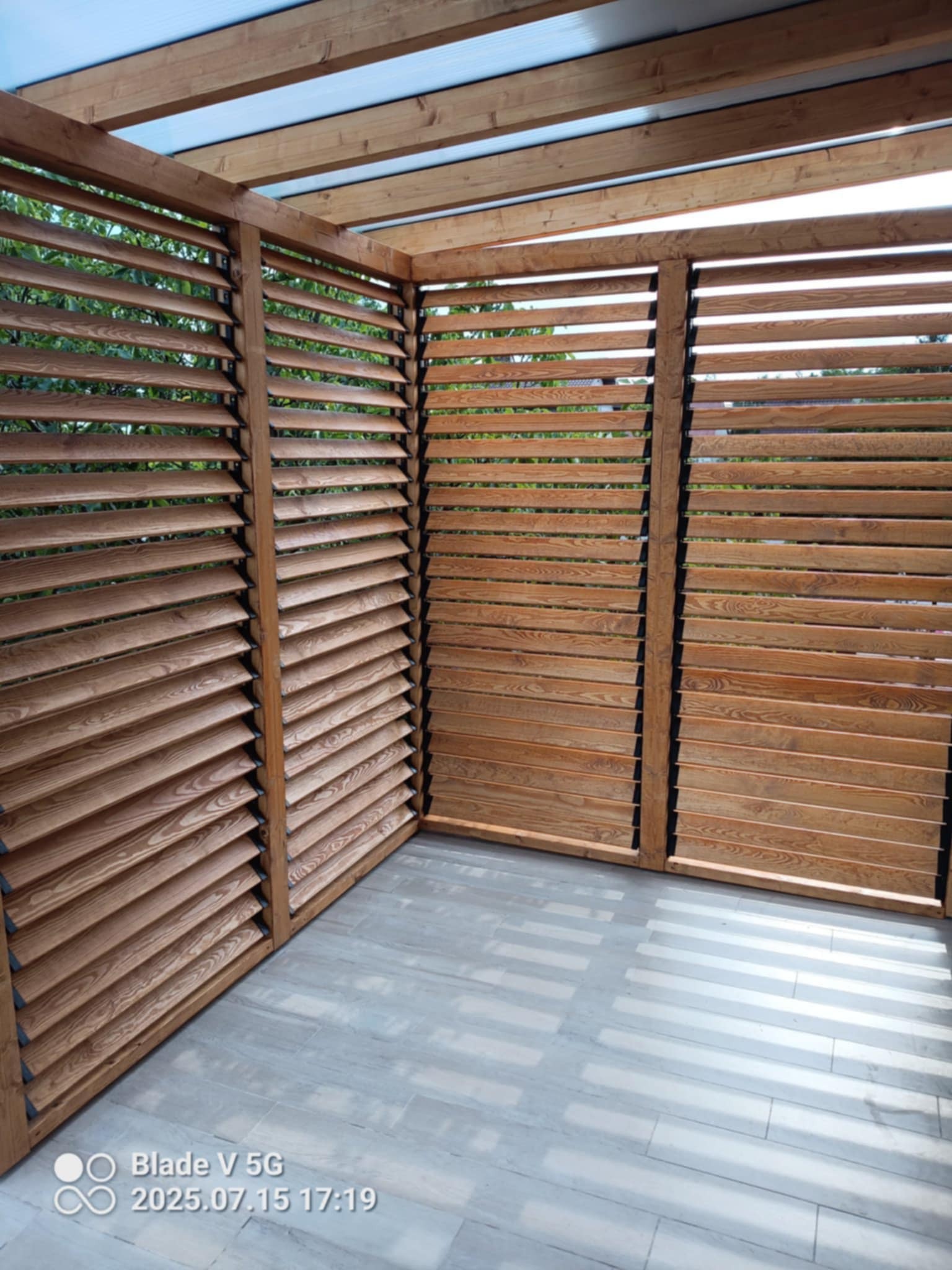 Drewniana pergola z lamelami i przezroczystym zadaszeniem, tworząca zacienioną przestrzeń na tarasie z szarymi płytkami. Widok z dołu, jasne oświetlenie.