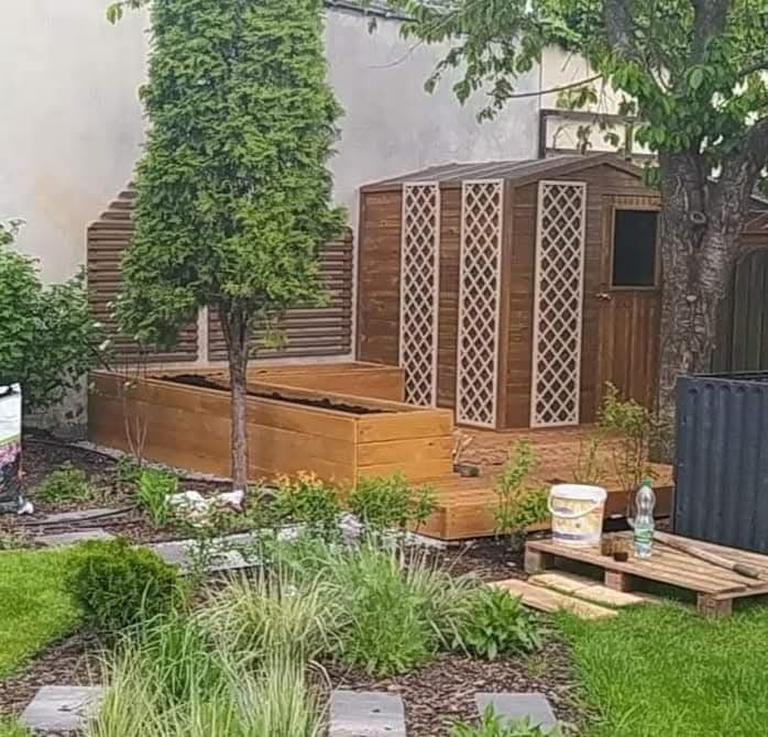 Drewniana konstrukcja ogrodowa z podwyższonymi grządkami, domkiem narzędziowym i tarasem, otoczona zielenią. Widoczne detale wykończeniowe i elementy dekoracyjne.
