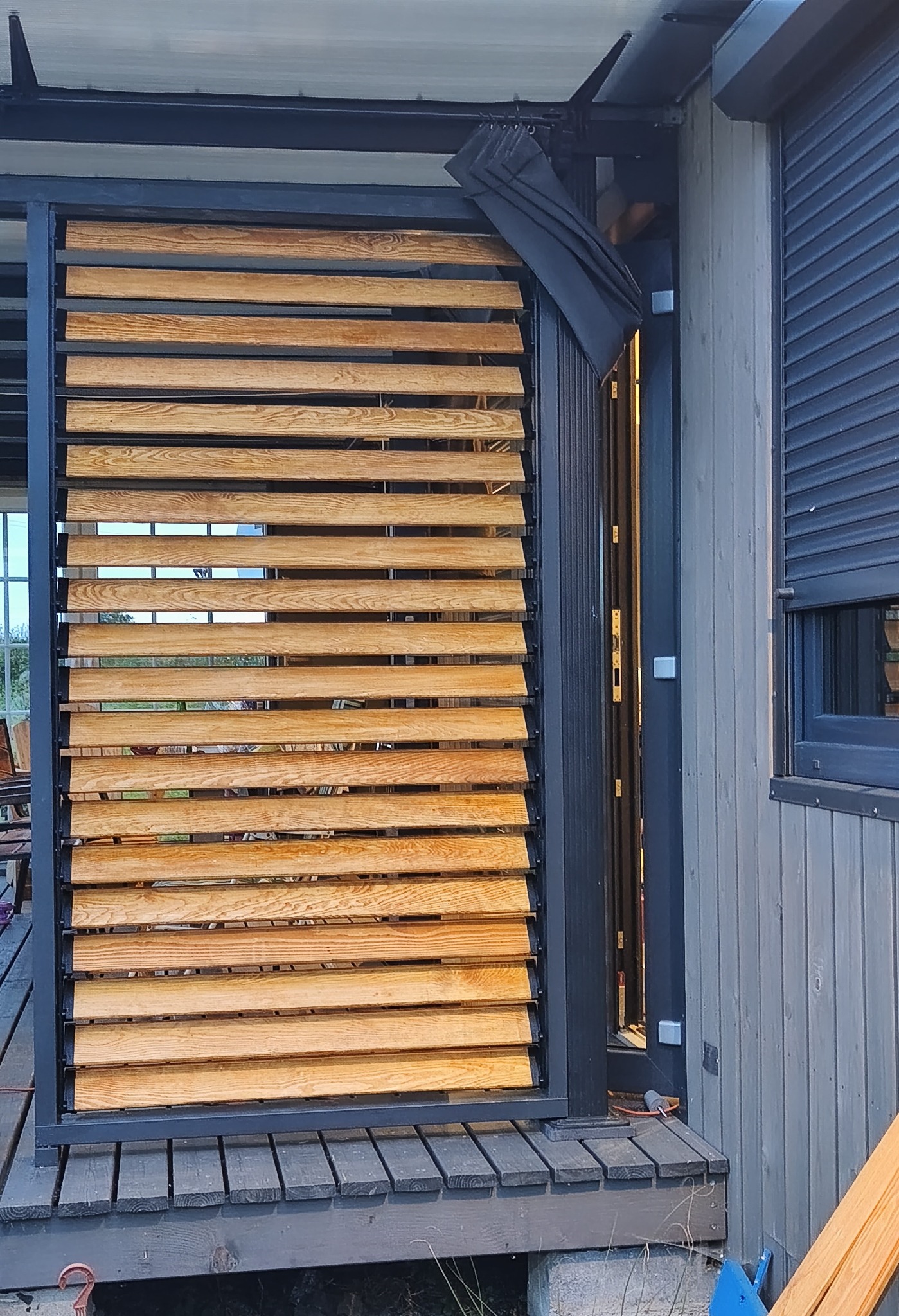 Nowoczesna pergola z drewnianymi lamelami i stalową konstrukcją, przylegająca do ściany budynku z szarą elewacją i roletami zewnętrznymi.