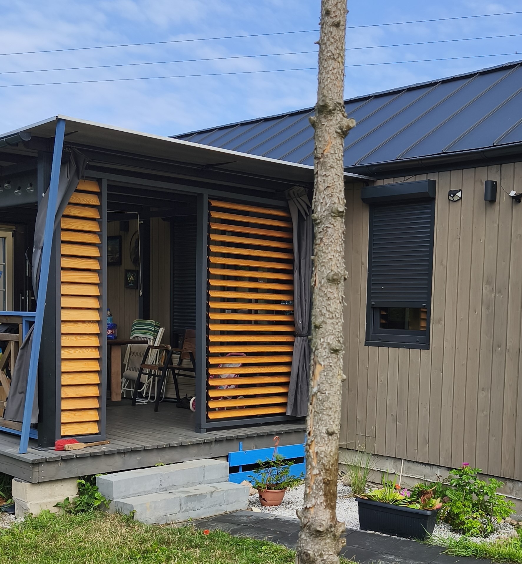 Nowoczesna, drewniana altana z przesuwnymi żaluzjami, szarą zasłoną i ciemnym dachem. Widoczne rośliny w donicach i fragment pnia drzewa na pierwszym planie.