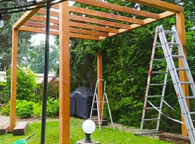 Drewniana pergola w trakcie budowy, z widocznymi elementami konstrukcyjnymi, drabiny i narzędzia na trawniku, w tle zielona roślinność.