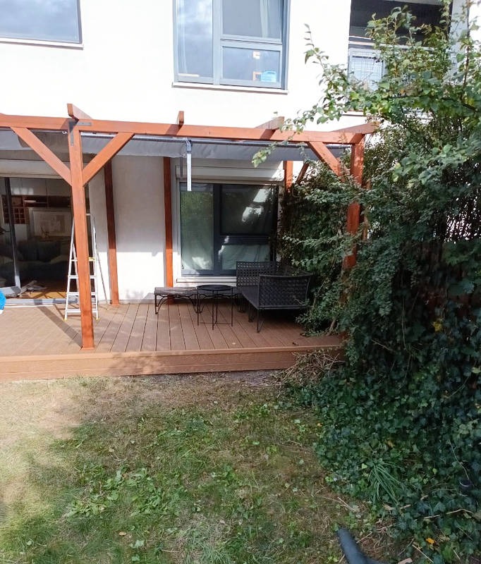 Drewniany taras z pergolą przy budynku. Na tarasie stolik kawowy i krzesła. Część tarasu z lewej zasłonięta, widoczna drabina. Zieleń w tle i na pierwszym planie.