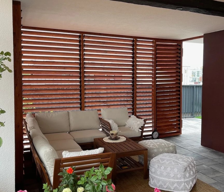 Zadaszony taras z drewnianymi żaluzjami w Warszawie. Wygodna sofa narożna, stolik kawowy i pufy tworzą przytulną strefę relaksu. Drewno w ciepłym odcieniu.