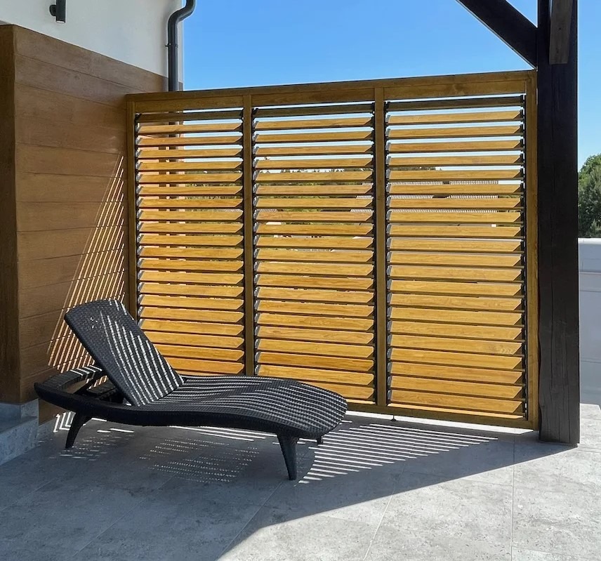 Drewniana osłona tarasu z poziomymi lamelami, rzucająca cienie na szary taras. Czarny leżak na pierwszym planie, błękitne niebo w tle.