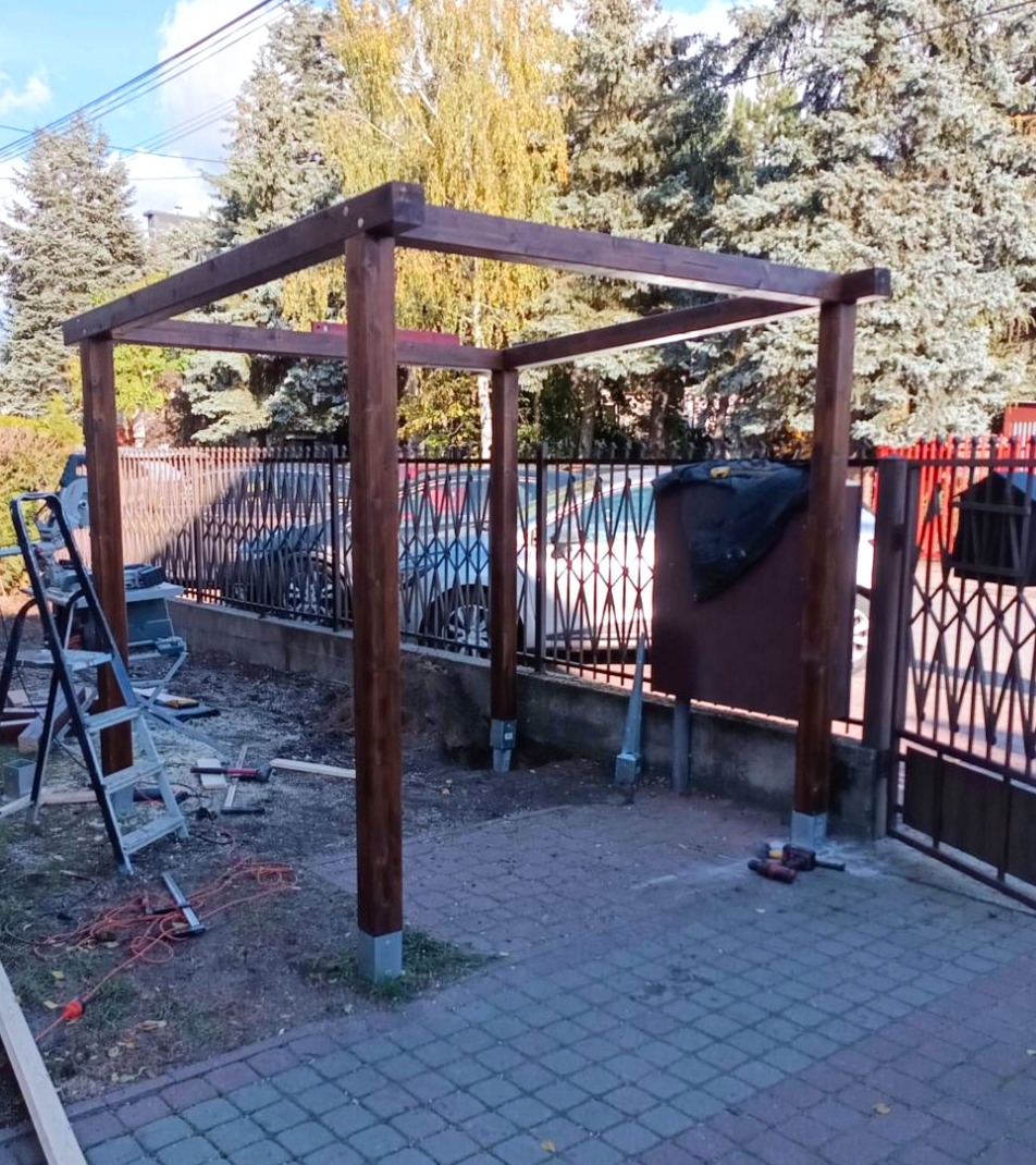 Drewniana konstrukcja pergoli w trakcie montażu, widoczne słupy i górna rama, narzędzia pracy obok drabiny, ogrodzenie w tle, kostka brukowa.