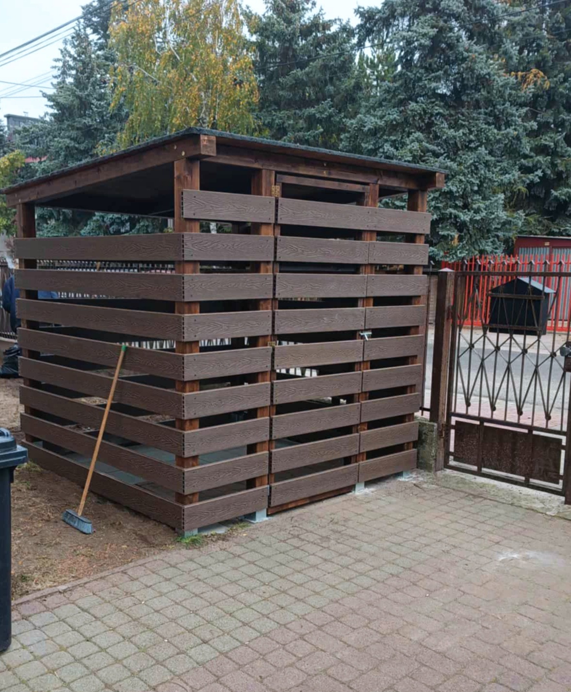 Drewniana konstrukcja z poziomych desek, prawdopodobnie osłona na śmietniki, z widoczną miotłą opartą o ścianę, na tle drzew i kostki brukowej.