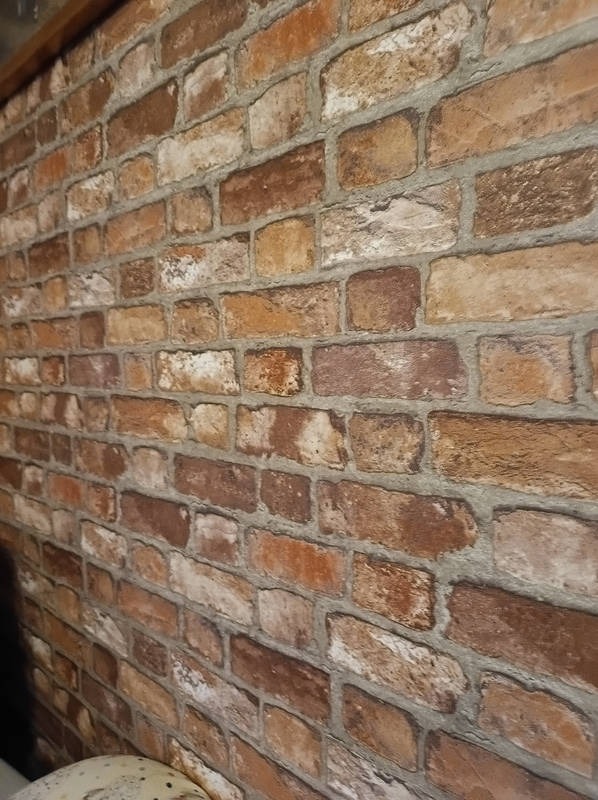 Ściana z cegieł w odcieniach brązu i beżu, z wyraźną fakturą i fugami. Ujęcie pod kątem, ukazujące perspektywę i głębię ściany. Widoczny fragment sufitu.