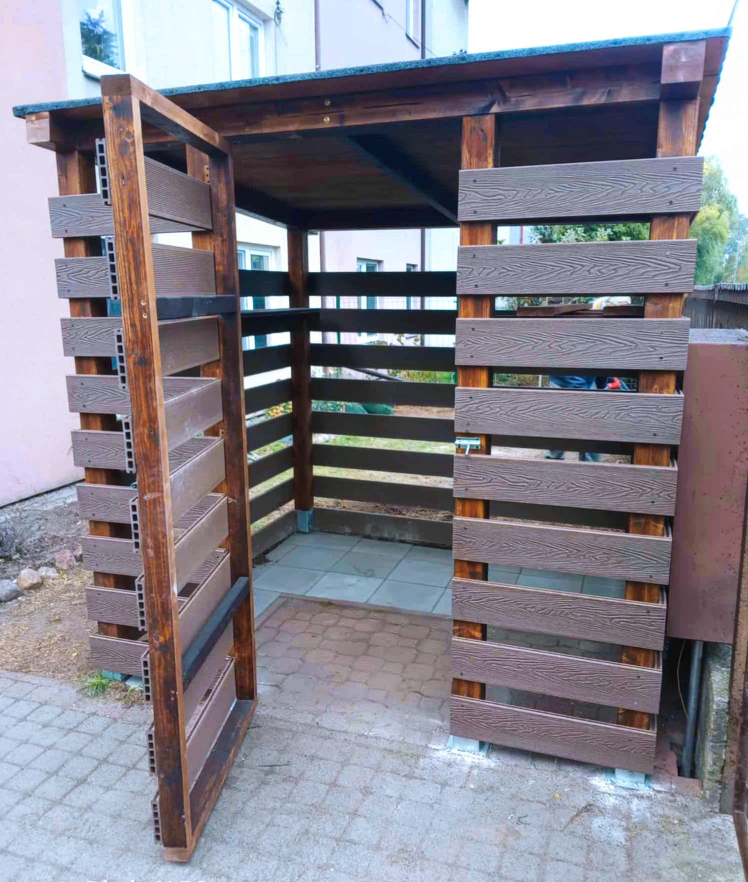 Drewniana pergola z ażurowymi ścianami z kompozytu, widok z zewnątrz. Otwarta boczna ściana. Podłoże z kostki brukowej. Widoczne elementy konstrukcyjne.