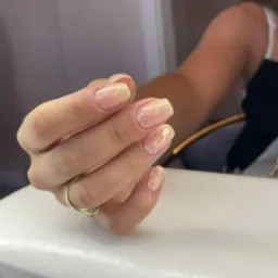 Dłoń z błyszczącym manicure hybrydowym w odcieniach beżu i złota, na serdecznym palcu złoty pierścionek. Efektowny, kwadratowy kształt paznokci.