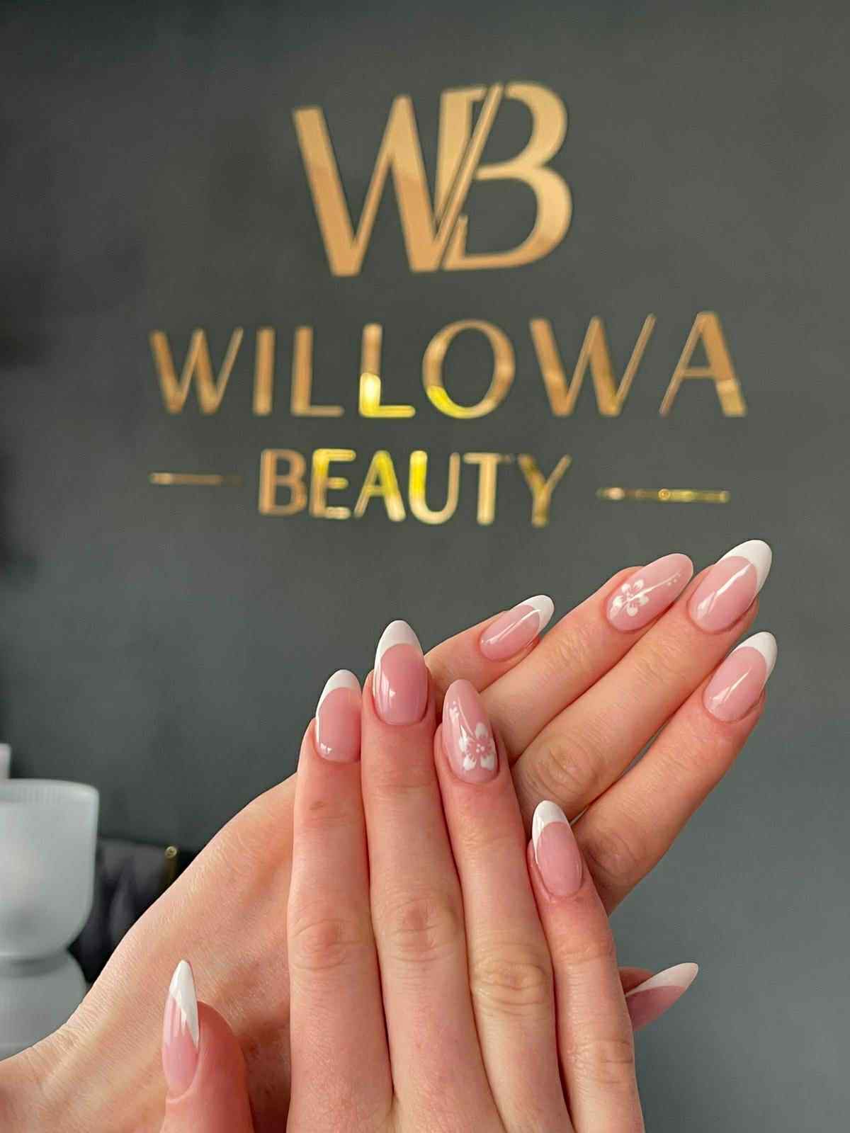 Dłonie z manicure francuskim i kwiatowym zdobieniem na tle ściany z logo 'Willow Beauty' w złotym kolorze. Delikatne, kobiece paznokcie w zbliżeniu.