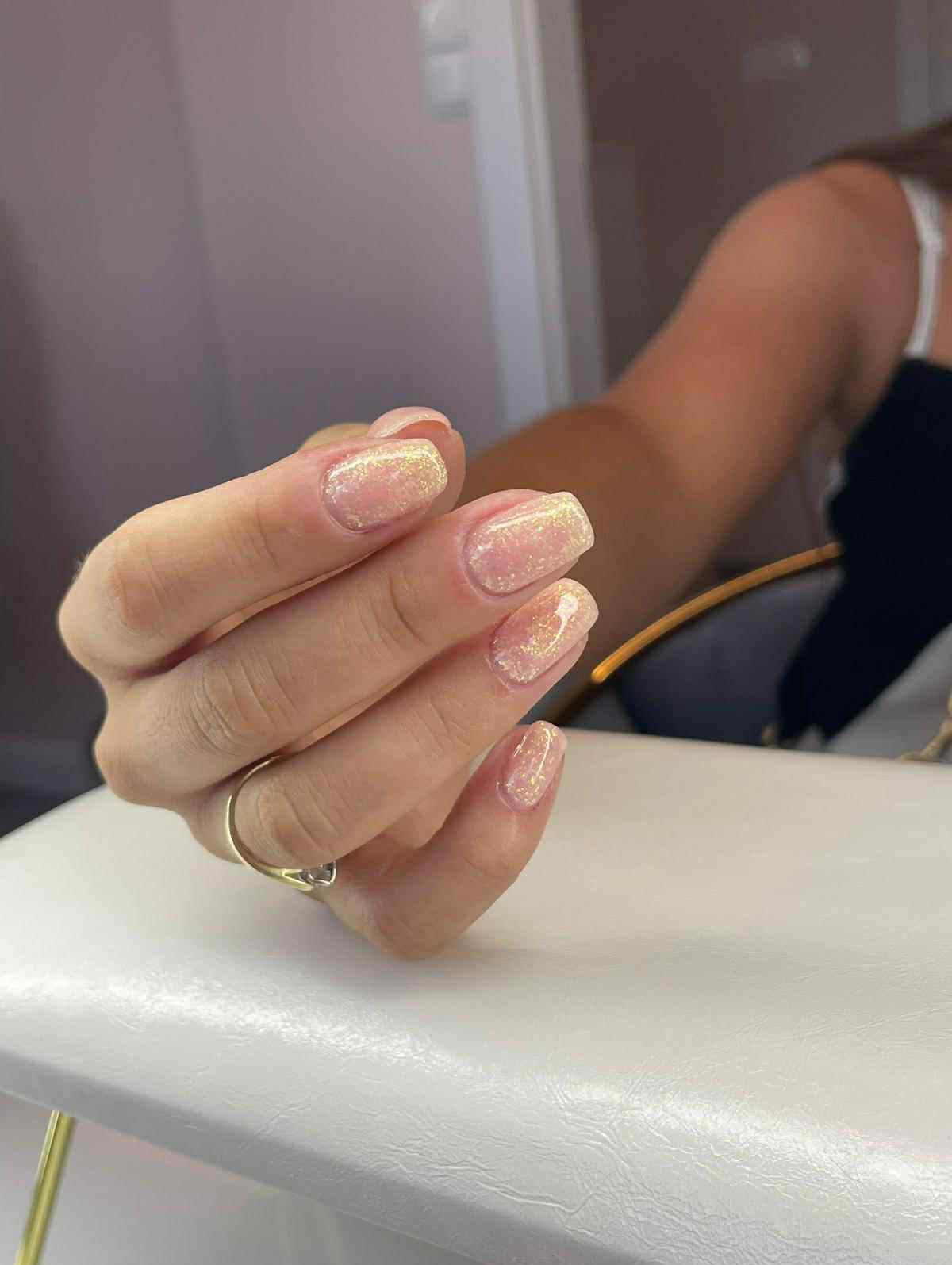 Dłoń z błyszczącym manicure hybrydowym w odcieniach beżu i złota, na serdecznym palcu złoty pierścionek. Efektowny, kwadratowy kształt paznokci.