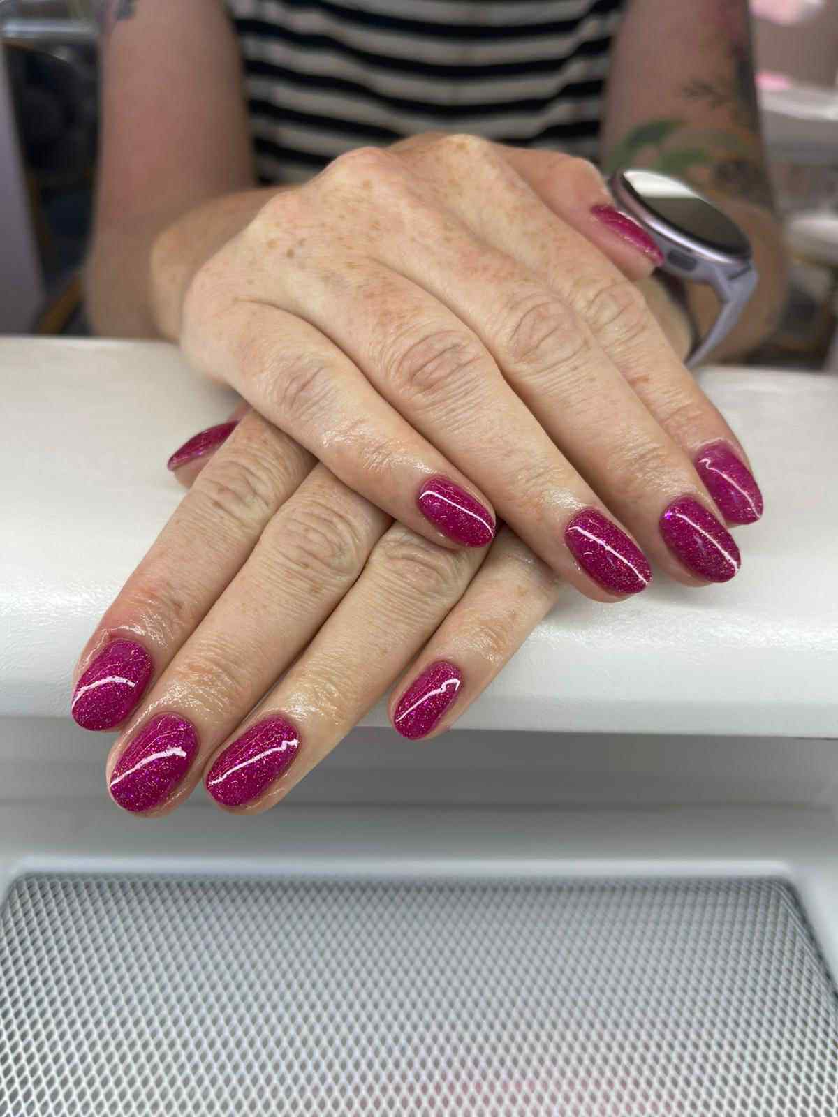 Dłonie z błyszczącym, malinowym manicure hybrydowym, widoczny zegarek na nadgarstku, wzorek w postaci białej linii na paznokciach, wykonany w salonie.