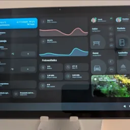 Tablet prezentuje interfejs systemu inteligentnego domu z danymi klimatycznymi, fotowoltaiką i sterowaniem urządzeniami w salonie, kuchni, łazience i garażu.