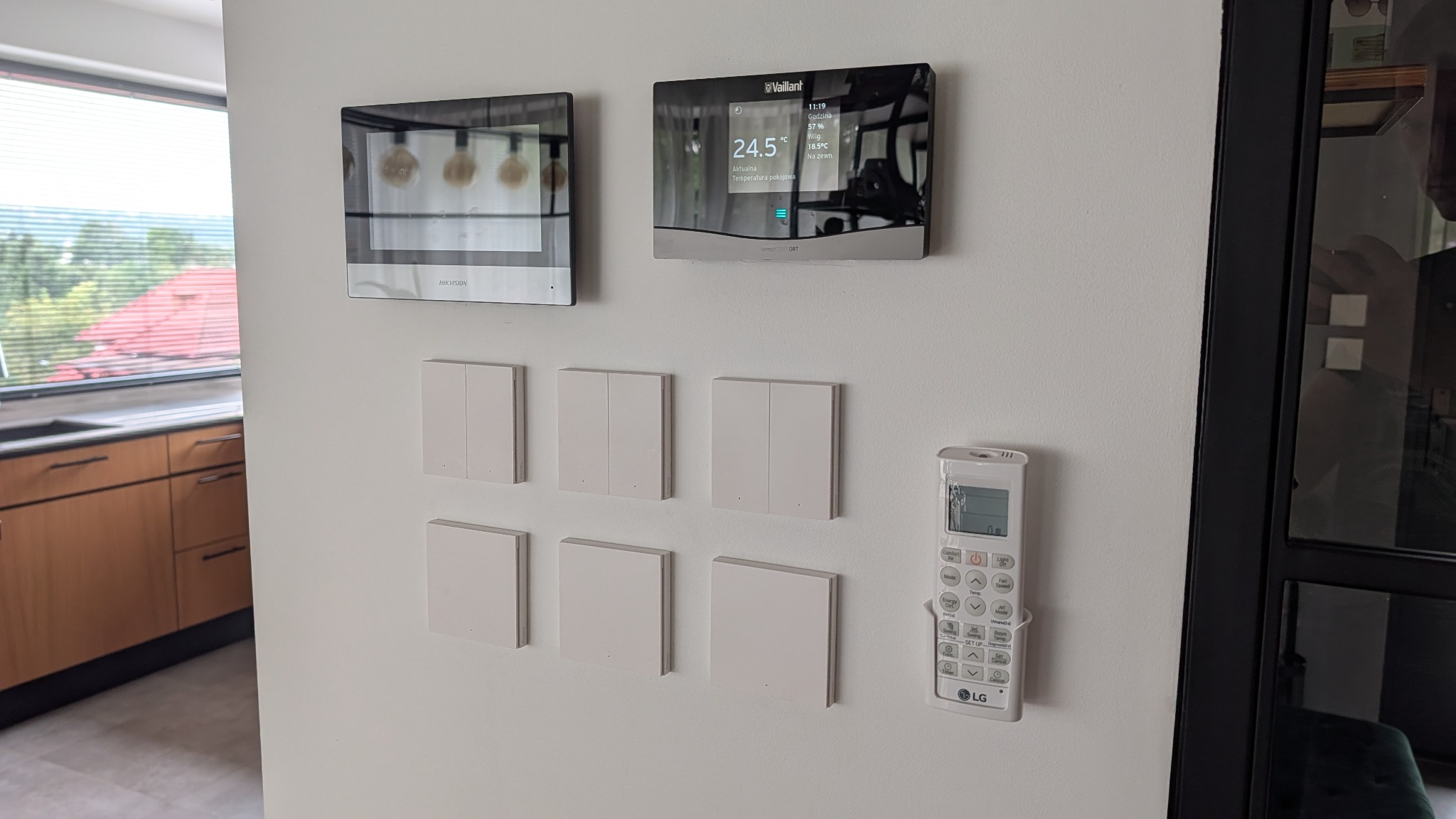 Panel sterowania inteligentnego domu z ekranem Vaillant, domofonem Hikvision, serią włączników i pilotem LG na białej ścianie. Widok na okno i fragment kuchni.