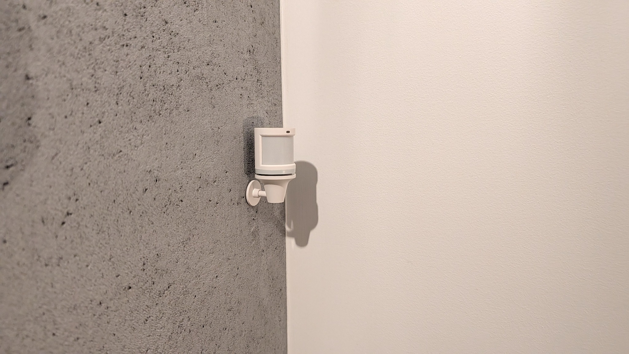 Czujnik ruchu zamontowany na szarej, teksturowanej ścianie w rogu pomieszczenia, obok gładkiej, białej ściany. Widoczny cień urządzenia.