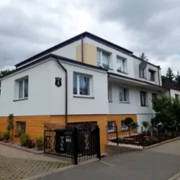 Dwukondygnacyjny budynek z białą elewacją i drewnopodobnym panelem na dole, widoczny z poziomu chodnika z kostki brukowej. Na ścianie herb.