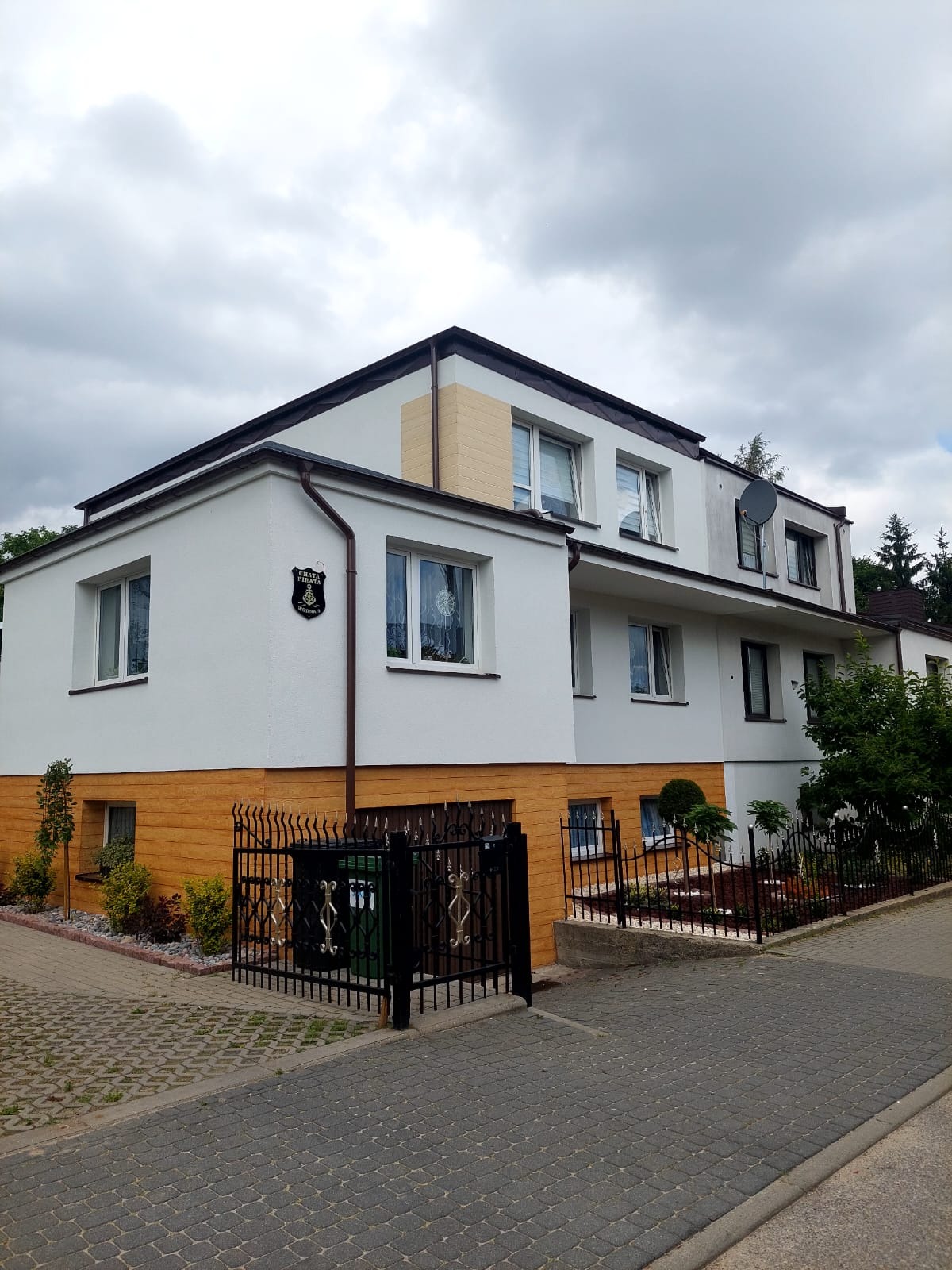 Dwukondygnacyjny budynek z białą elewacją i drewnopodobnym panelem na dole, widoczny z poziomu chodnika z kostki brukowej. Na ścianie herb.