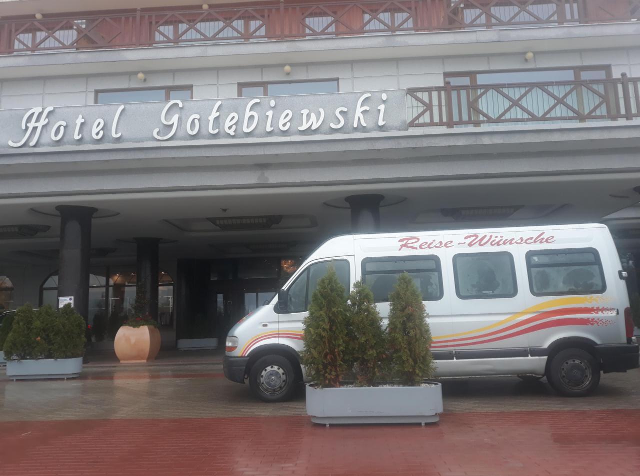 Biały bus z napisem 'Reise-Wünsche' zaparkowany przed hotelem Gołębiewski, widoczne rośliny w donicach i mokra, czerwona kostka brukowa.