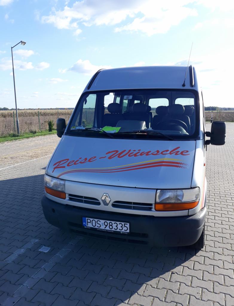 Srebrny bus Renault z napisem 'Reise-Wünsche' na masce, zaparkowany na brukowanym placu, widoczna polska tablica rejestracyjna POS 98335, antena na dachu i pole w tle.