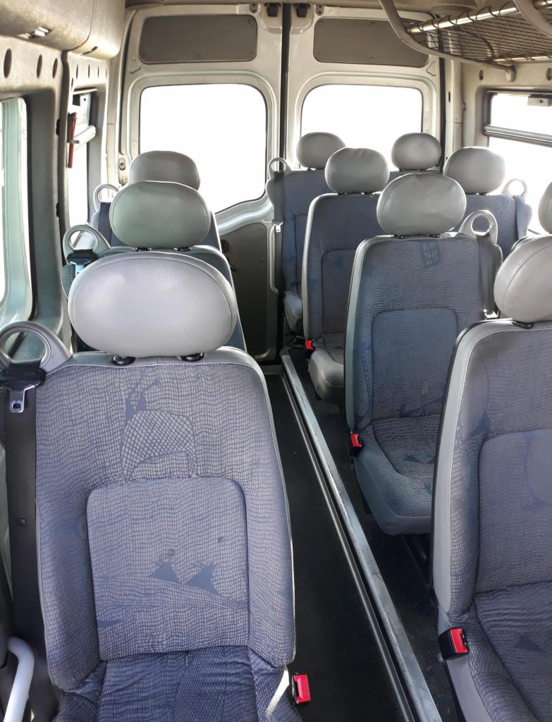 Wnętrze busa z rzędami szarych foteli z zagłówkami, widok z tyłu pojazdu w kierunku jasnych drzwi.