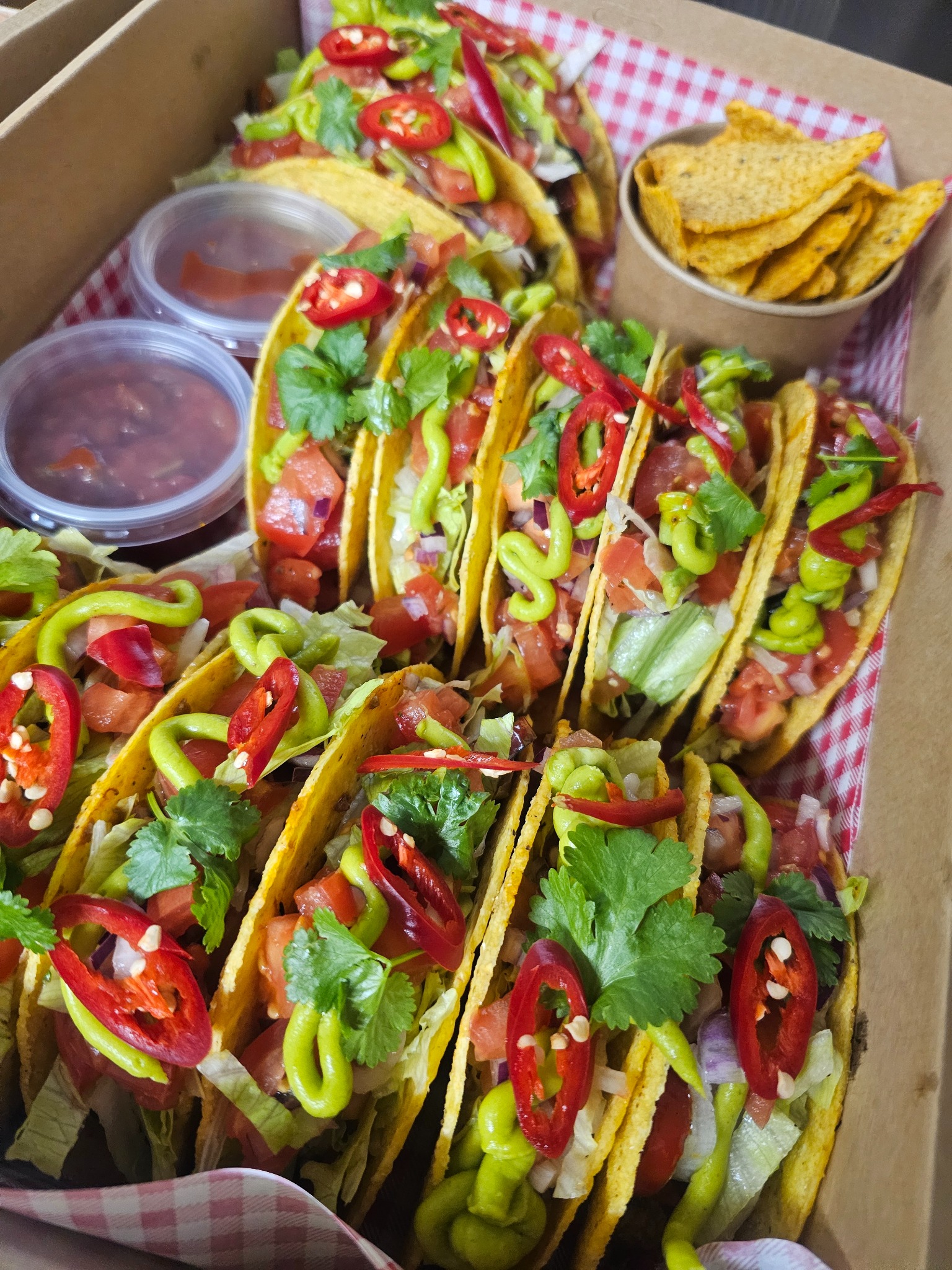 Tacos z warzywami i sosem w kartonowym pudełku na czerwono-białej serwetce, uzupełnione nachosami i dodatkowymi sosami.