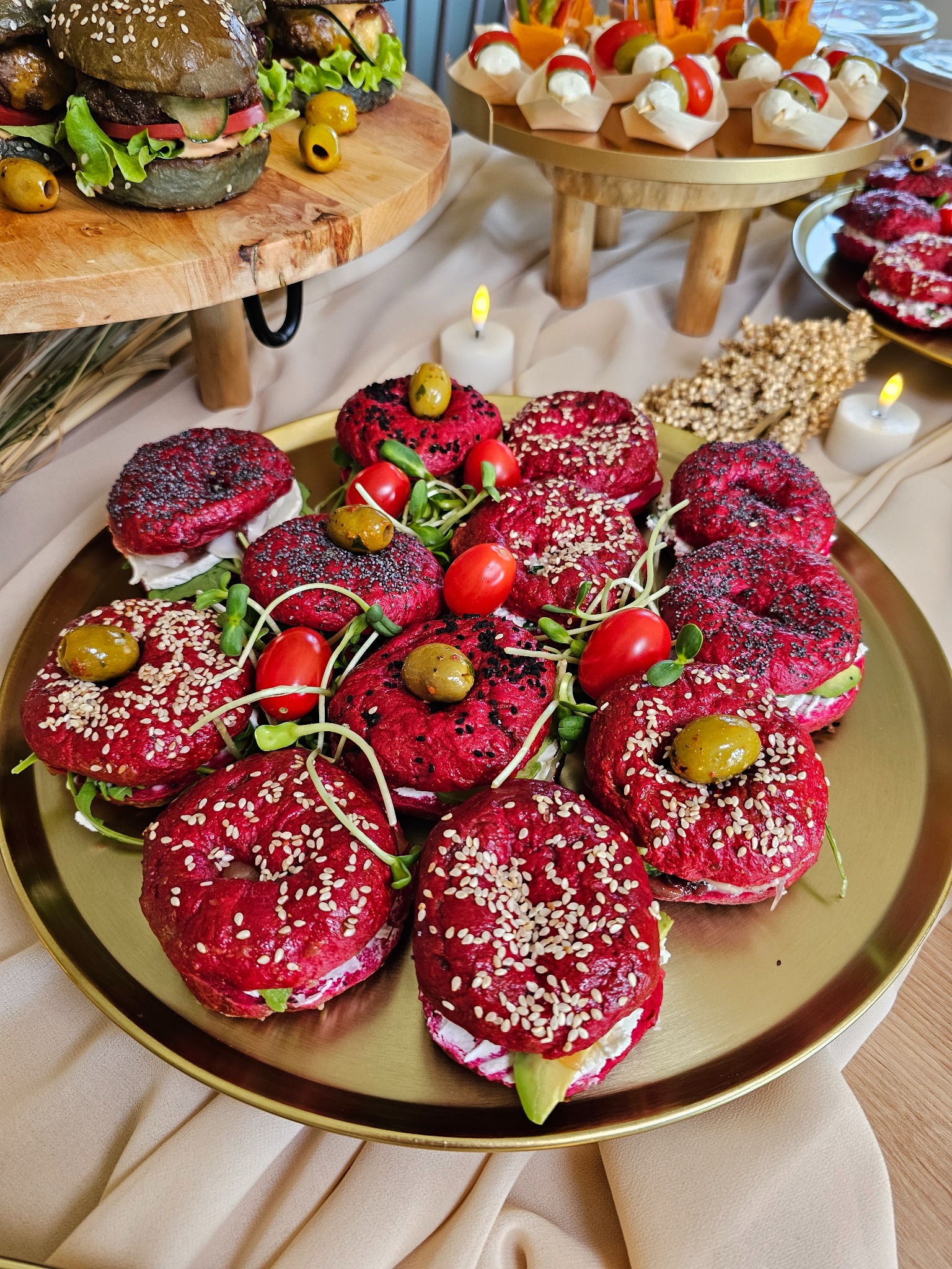 Złota taca z czerwonymi mini-burgerami, posypanymi sezamem i makiem, udekorowanymi oliwkami i pomidorkami koktajlowymi. W tle czarne burgery i przekąski.