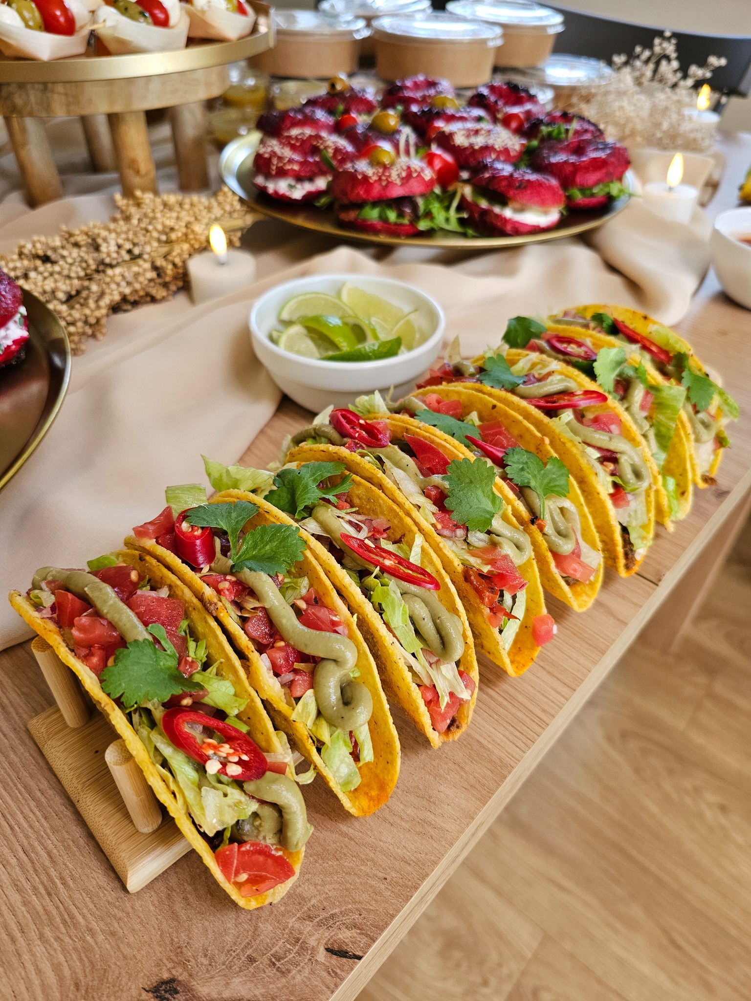 Rząd apetycznych tacos w drewnianym stojaku, obok miseczka z limonkami i talerz z mini burgerami na stole, dekoracja świecami i suszem. Elegancki catering.