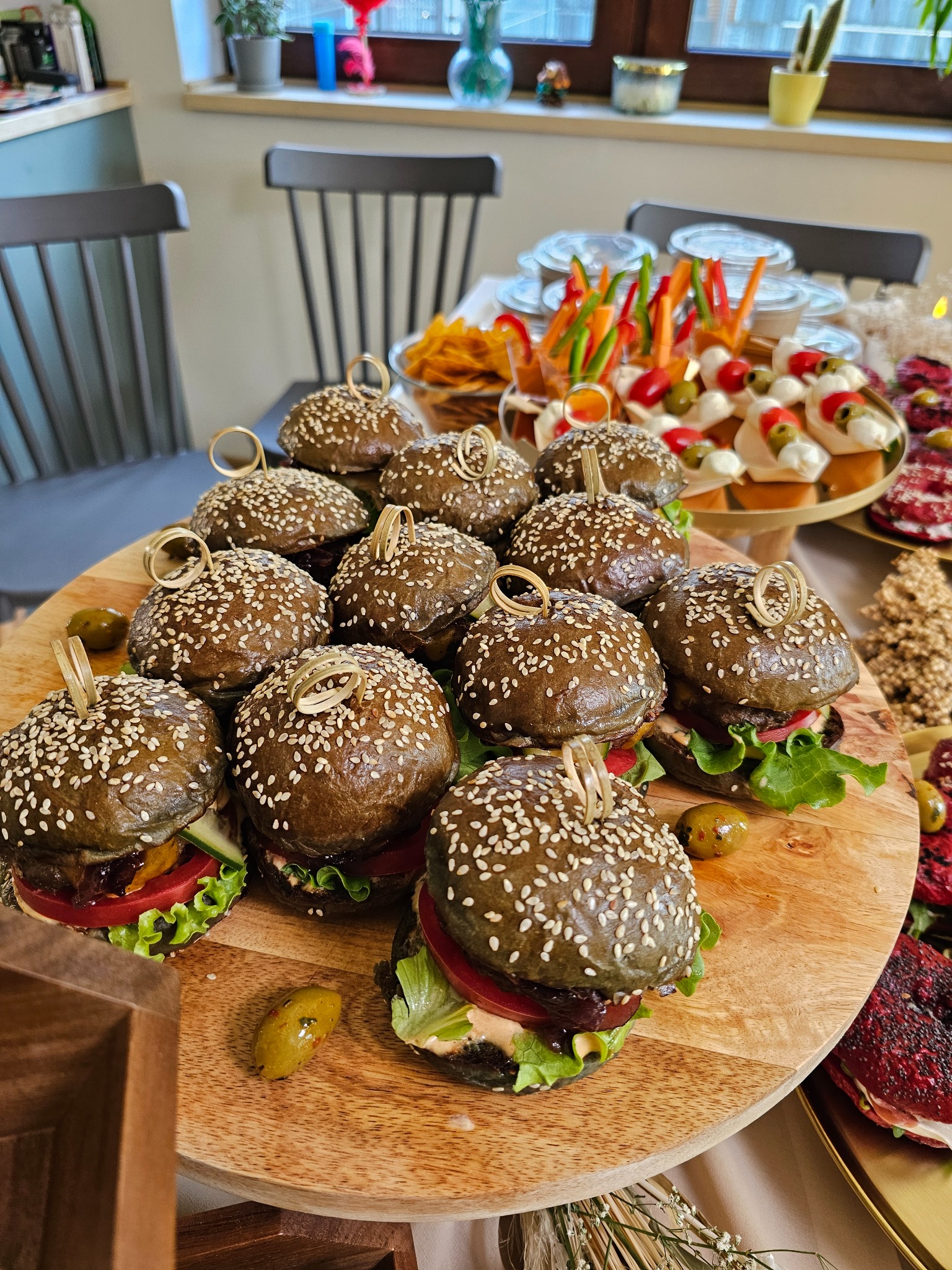 Ułożone na drewnianej desce burgery z czarną bułką i sezamem, obok przekąski warzywne i mini-mozzarella. Elegancki catering na przyjęcie.