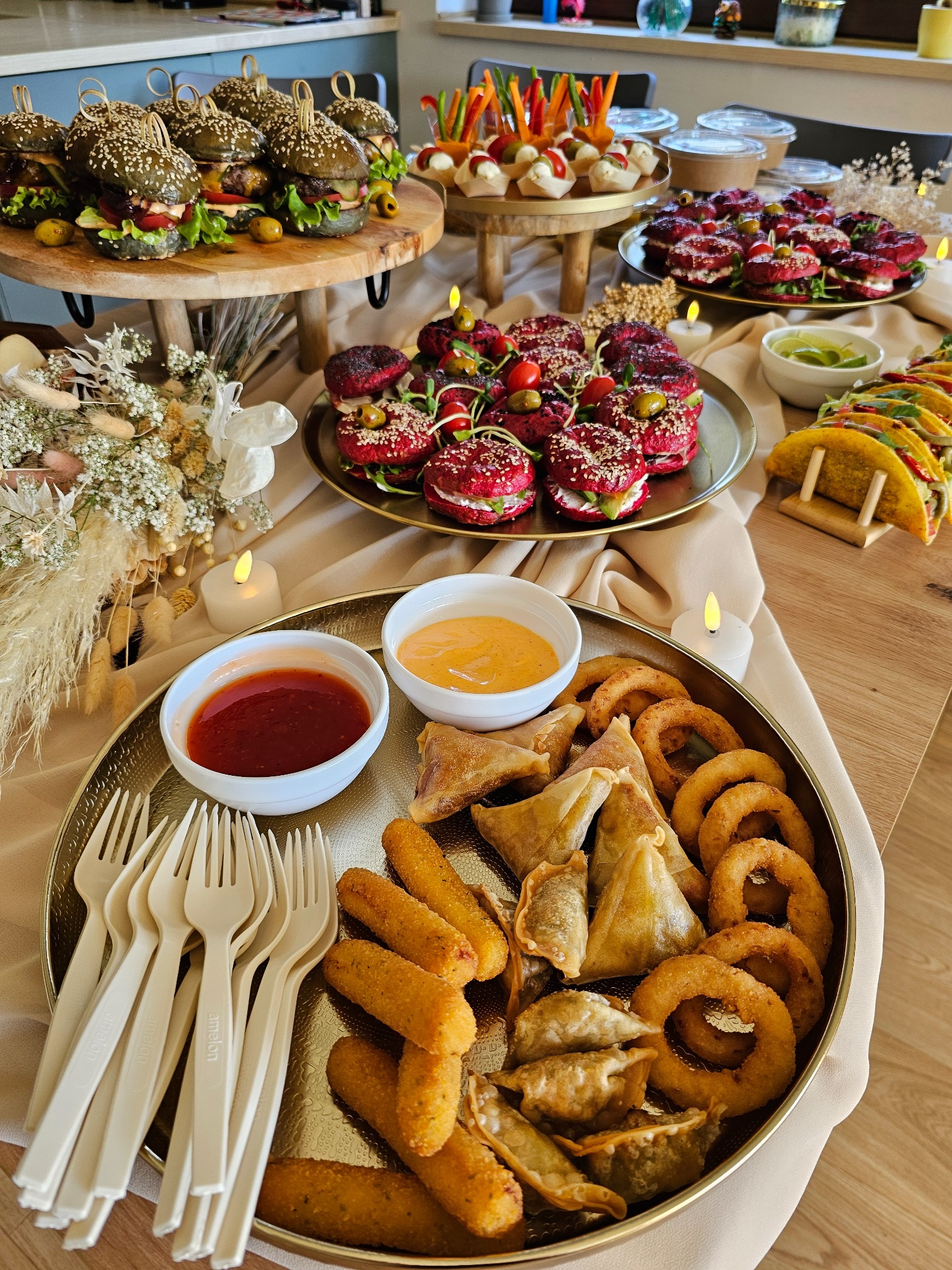 Bogato zastawiony stół z różnorodnymi przekąskami: kolorowe burgery, tacos, pierożki, krążki cebulowe, sosy i bukiet kwiatów. Elegancki catering na przyjęcie.