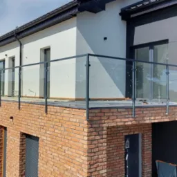 Nowoczesna balustrada szklana z ciemnymi profilami na balkonie budynku z cegły. Widok z zewnątrz, jasne tło, częściowo widoczne okna i elewacja.