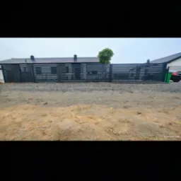 Nowoczesna, pozioma brama wjazdowa z metalowymi panelami w kolorze antracytowym, zintegrowana z ogrodzeniem. Widok z zewnątrz, na tle domu z ciemnym dachem.