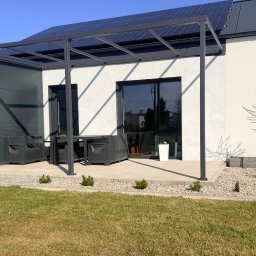 Metallum Adrian Pawlak - Nowoczesna, grafitowa pergola z metalową konstrukcją, zadaszająca taras z meblami ogrodowymi. Na dachu panele słoneczne, w tle biała elewacja domu.