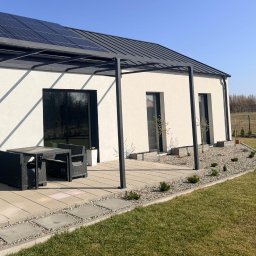 Metallum Adrian Pawlak - Nowoczesna pergola z ciemną ramą, przylegająca do białego domu z panelami słonecznymi. Na tarasie zestaw mebli ogrodowych. Minimalistyczny design, słoneczny dzień.