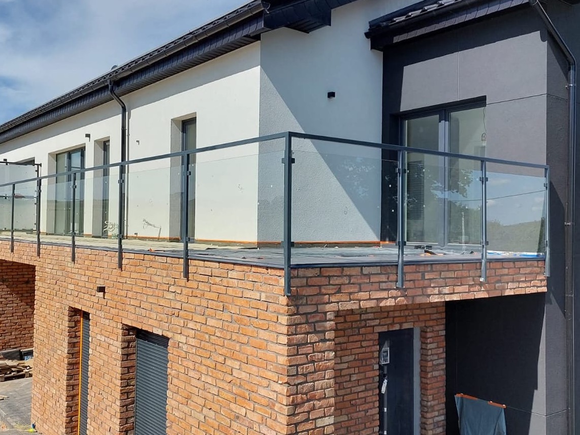 Nowoczesna balustrada szklana z ciemnymi profilami na balkonie budynku z cegły. Widok z zewnątrz, jasne tło, częściowo widoczne okna i elewacja.