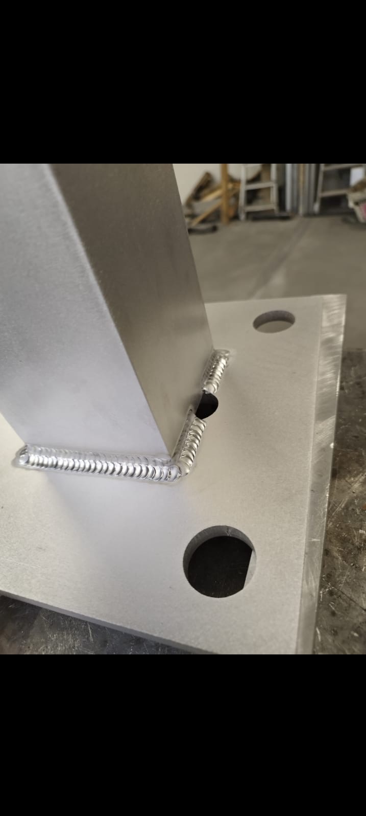 Szczegółowe zbliżenie na spawany element metalowy, widoczne precyzyjne spawy łączące pionowy profil z poziomą płytą. Widoczne otwory w płycie.