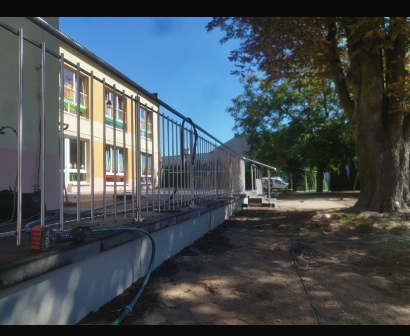 Nierdzewna balustrada przy budynku, widok z boku, narzędzia na pierwszym planie. Metalowe słupki i poziome rury tworzą barierę. W tle drzewa i błękitne niebo.