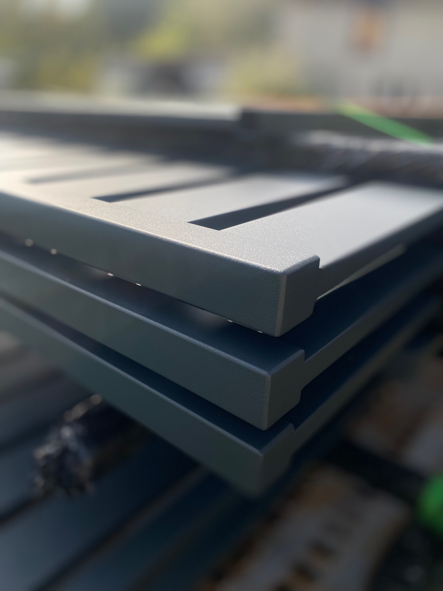 Stos szarych, metalowych przęseł ogrodzeniowych z prostokątnymi wycięciami, ułożonych jeden na drugim w słoneczny dzień. Widoczna faktura proszkowego malowania.