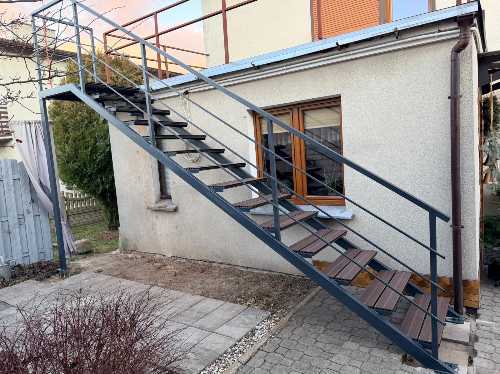 Zewnętrzne schody metalowe z drewnopodobnymi stopniami, prowadzące na balkon. Szara konstrukcja na tle jasnego budynku z oknami. Widok z dołu, z boku.