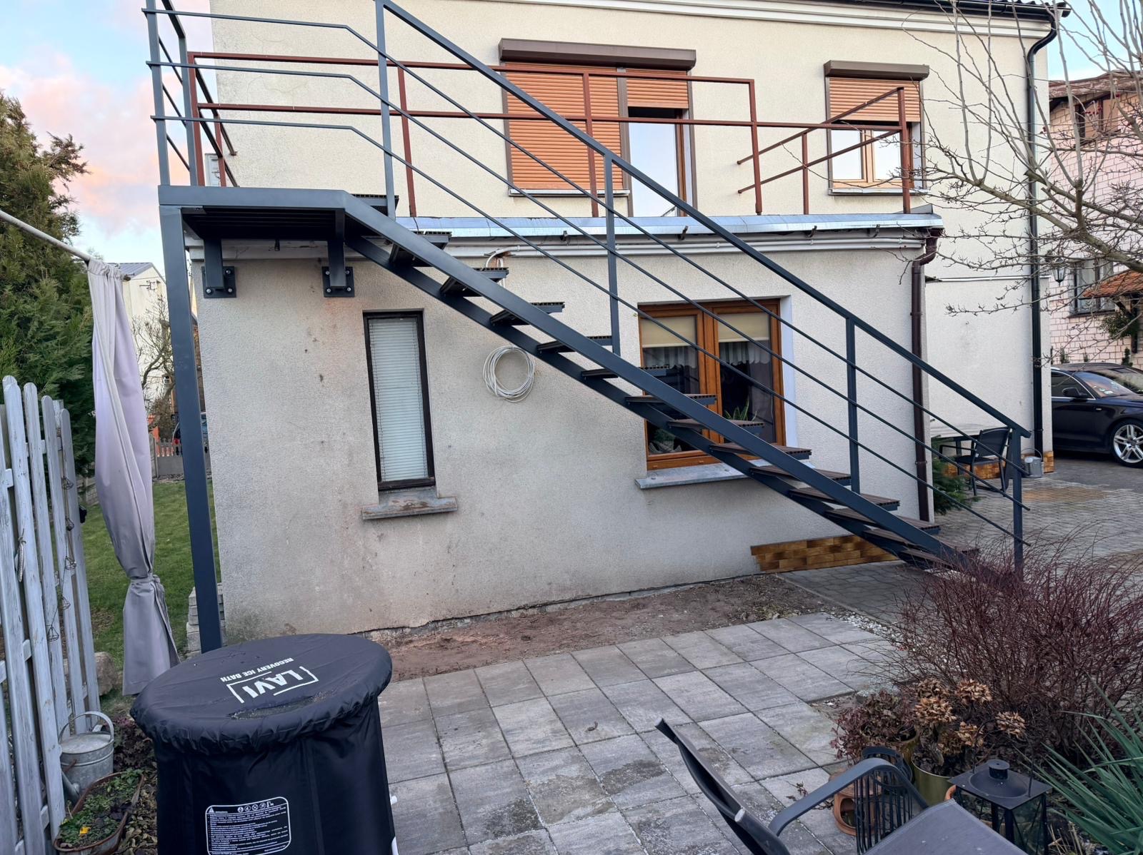 Zewnętrzne schody metalowe z balustradą prowadzące na balkon z brązowymi roletami, zamontowane przy jasnym budynku. Widok z dołu na szarej kostce brukowej.