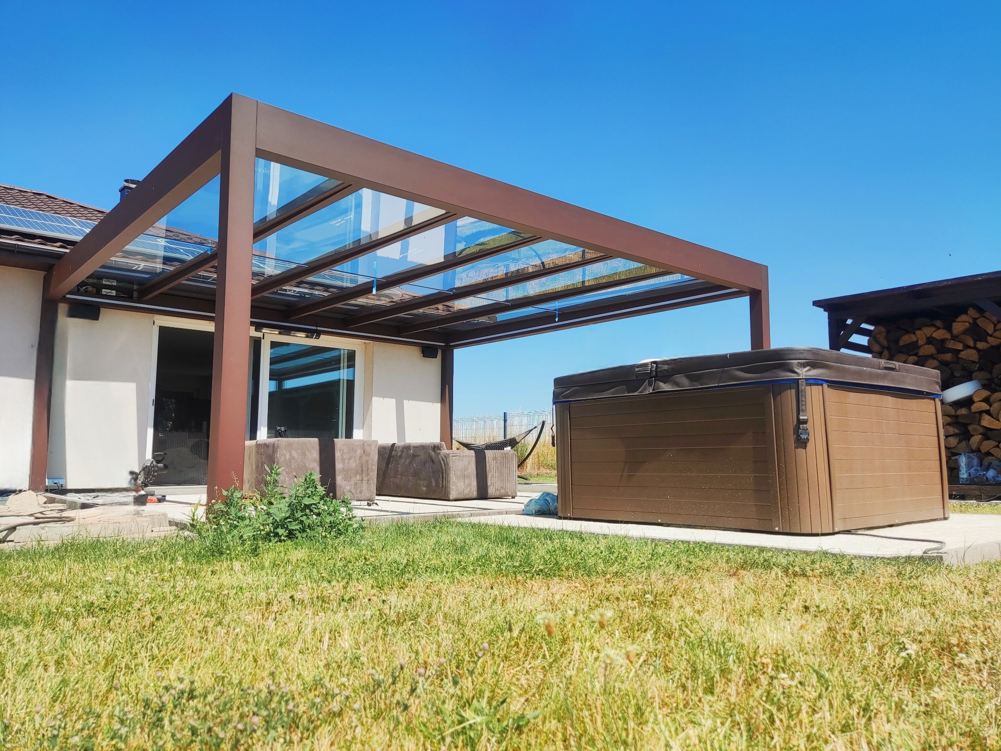 Brązowa pergola z przeszklonym dachem przylegająca do domu. Obok jacuzzi, trawnik i narzędzia budowlane. Słoneczny dzień.