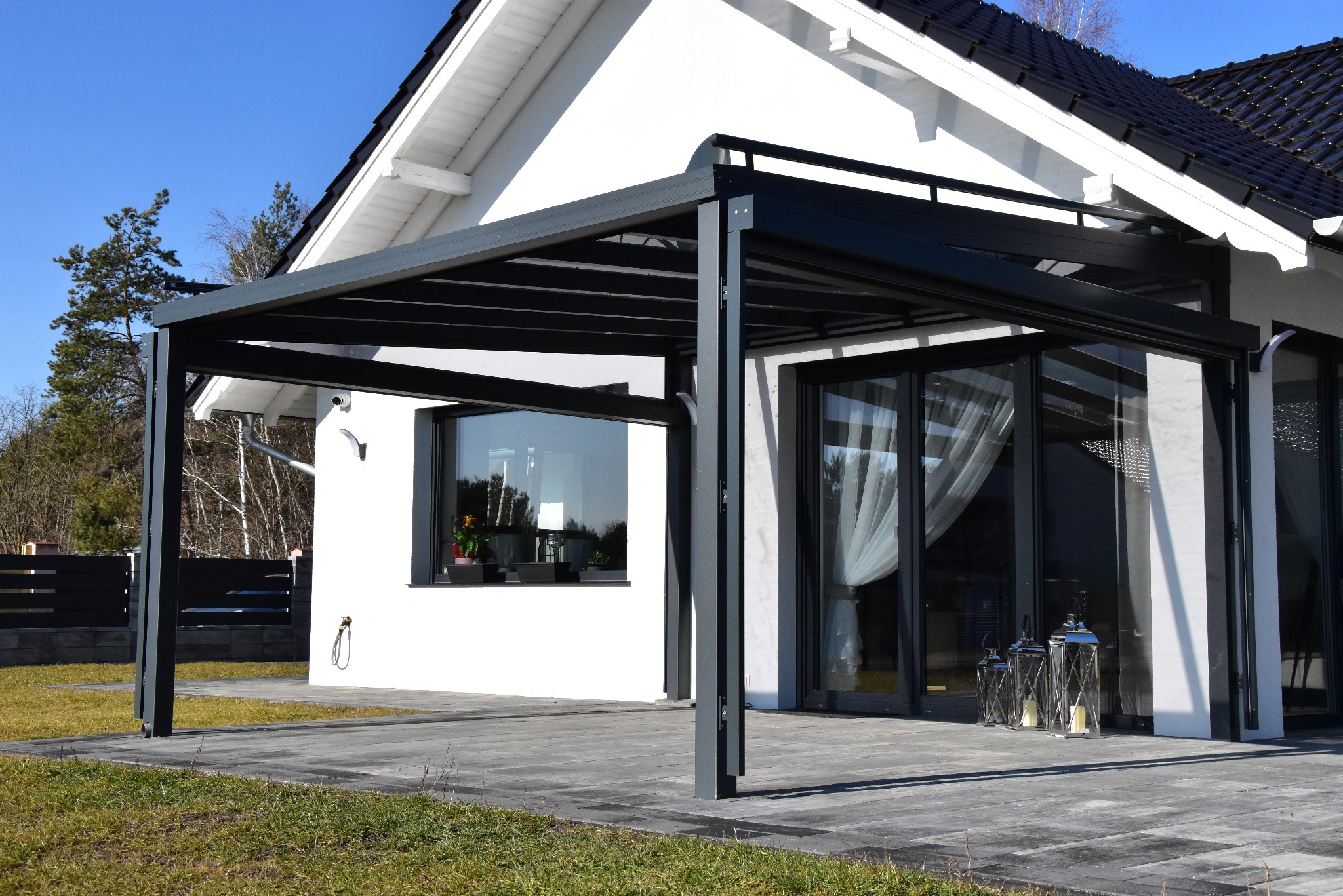 Nowoczesna, grafitowa pergola metalowa przy białym domu z dużymi oknami, oświetlona słońcem. Na tarasie stoją dekoracyjne lampiony.