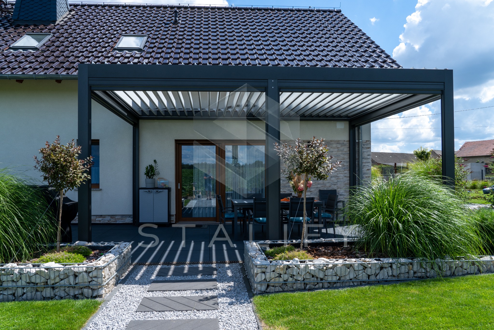 Nowoczesna, grafitowa pergola metalowa z regulowanymi lamelami, przylegająca do jasnego domu. Widoczne elementy dekoracyjne i zadbany ogród.